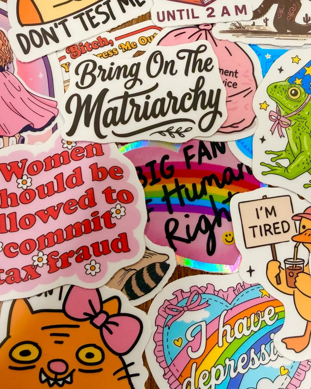 A dense collage of colorful die-cut stickers featuring humorous, political, and quirky cartoon illustrations and text slogans, overlapping each other across the full frame.